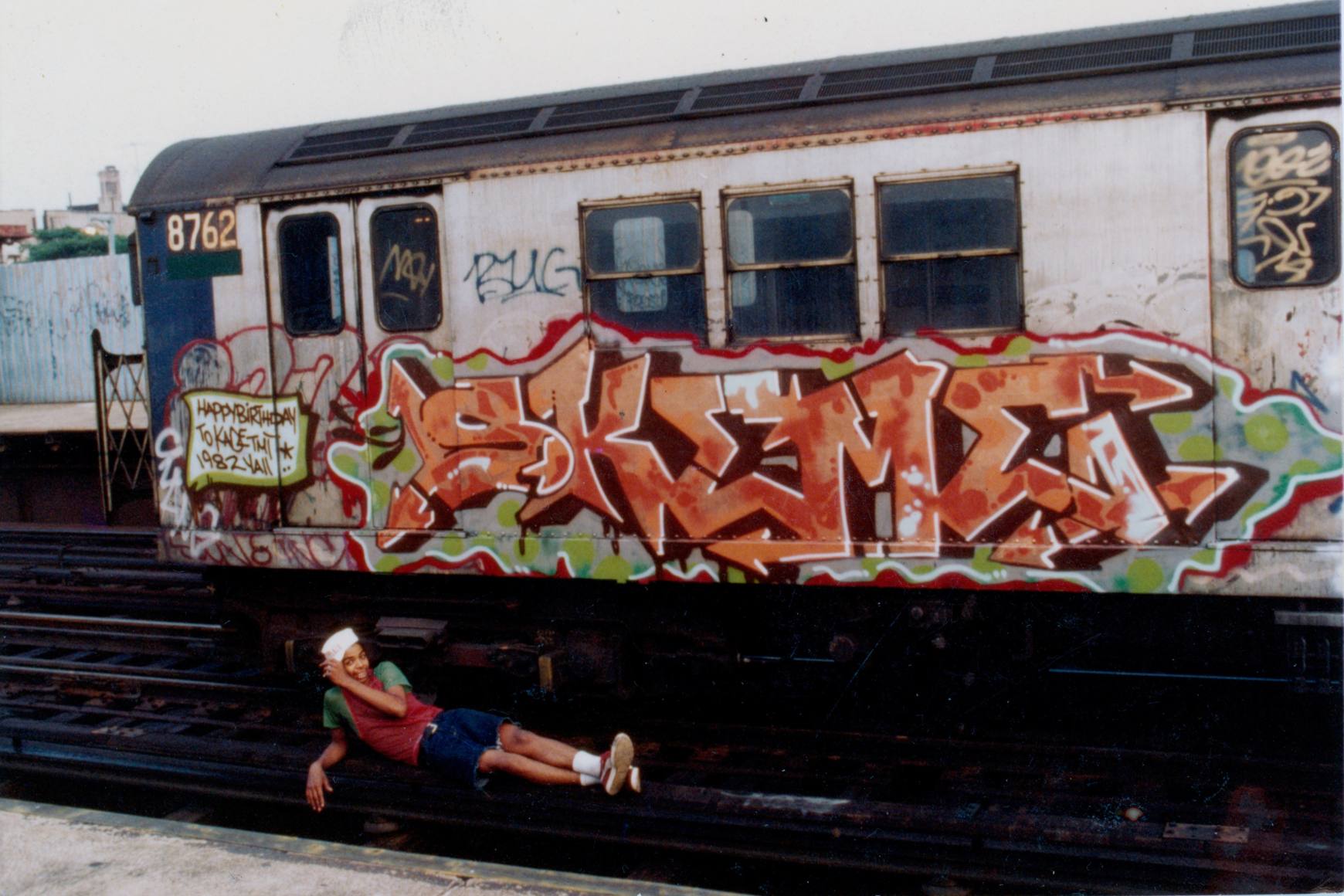 Skeme lies on the subway track below a parked subway car with a large orange artwork by him splashed across it. 